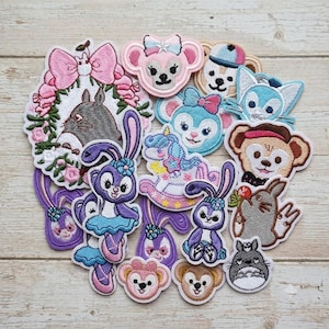 May include: A collection of embroidered fabric patches featuring various cartoon characters, including a gray forest creature in a floral wreath, purple and blue rabbits, a unicorn on a rocking horse, and several small animal faces in pastel colors.