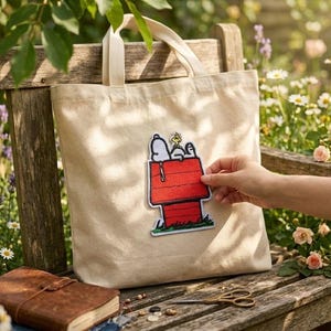 Puede incluir: Un bolso de tela beige con un gran parche bordado del personaje de dibujos animados Snoopy tumbado sobre una caseta de perro roja con Woodstock posado en el tejado. El parche está centrado en la parte delantera del bolso de dos asas.