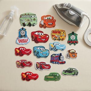 Könnte beinhalten: Eine Sammlung bestickter Stoffaufnäher mit verschiedenen Zeichentrickfahrzeugen und Zügen, darunter Figuren aus Thomas & Friends und Cars, angeordnet neben einer transparenten Sprühflasche und einem weißen elektrischen Bügeleisen.