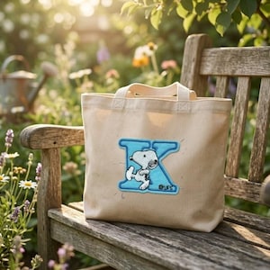 May include: A beige canvas tote bag featuring a large, light blue embroidered letter K with a white Snoopy character graphic positioned in the center. The bag has two top handles and a rectangular shape, displaying a clear, stitched design.