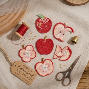 Könnte beinhalten: Ein Set aus gestickten Apfel-Aufnähern mit einem ganzen roten Apfel, einem aufgeschnittenen Apfel und Apfelscheiben mit Blütenmotiven. Das Set enthält eine Spule mit rotem Garn, einen goldenen Fingerhut und eine Schere mit einem Papieranhänger.