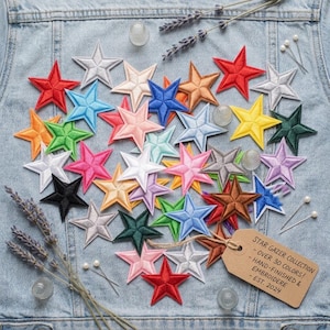 Puede incluir: Una colección de más de 30 parches bordados en forma de estrella de múltiples colores sobre un fondo de mezclilla. Se adjunta una etiqueta de papel marrón que dice: STAR GAZER COLLECTION - OVER 30 COLORS! - HAND-FINISHED & EMBROIDERED - EST. 2024.