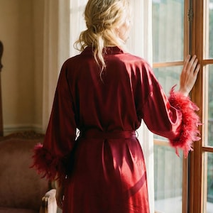 May include: A burgundy satin robe with feather trim on the sleeves. The robe is tied at the waist with a matching belt. A woman is standing by a window, wearing the robe. A large mirror and chair are in the background.