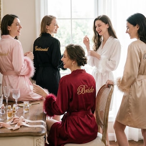 Può includere: Cinque donne in vestaglie di seta, alcune con finiture in piume, sono riunite in una stanza ben illuminata. Gli abiti sono in tonalità di rosa, nero, bianco e bordeaux. Ricami dorati sugli abiti recitano "Bride" e "Bridesmaid". La scena suggerisce un addio al nubilato.