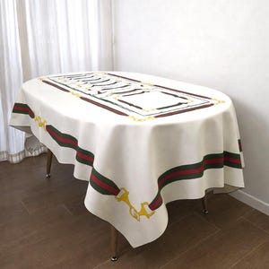 May include: Oval white tablecloth with a decorative border featuring a red and green stripe design. The tablecloth has a central rectangular pattern with black and gold accents, and is draped over a wooden table.