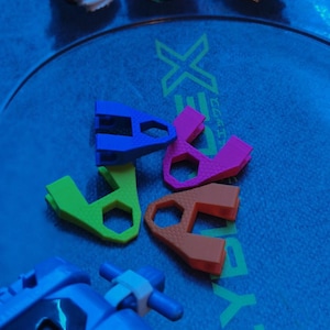 May include: Three Beyblade spinning tops in white, green, and dark metallic colors, accompanied by four plastic tools in blue, pink, green, and orange, and a blue launcher, all resting on a circular Beyblade X arena with green trim.