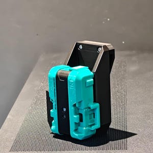 Könnte beinhalten: Ein schwarzer Beyblade X-Starterhalter aus Kunststoff mit einem türkisfarbenen Starter an der Vorderseite. Der Halter hat ein vertikales Design mit sichtbaren Schrauben und einer strukturierten Oberfläche, die das mechanische Spielzeugteil sicher aufnimmt.