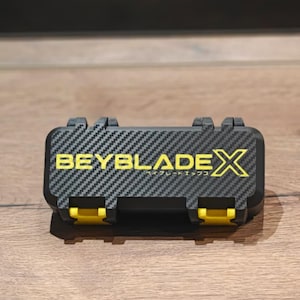 Op de afbeelding: Een rechthoekige zwarte opbergkoffer met een koolstofvezel textuurpatroon. De voorzijde is voorzien van de gele tekst BEYBLADEX met Japanse karakters eronder. De koffer heeft twee felgele sluitingen aan de onderkant voor een veilige sluiting.