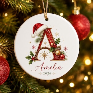 May include: A round white ceramic Christmas ornament featuring a large red letter A decorated with holly leaves, red berries, gold baubles, and snowflakes. A Santa hat sits atop the letter, and the name Amelia and the year 2026 are printed below.