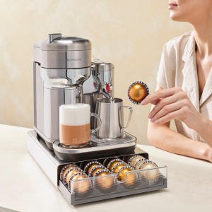May include: A stainless steel Nespresso coffee machine with a milk frother, a small stainless steel pitcher, and a glass mug with a latte. A hand holds a coffee pod. A drawer below holds more pods.