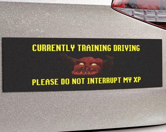 May include: A rectangular black bumper sticker with yellow text that reads CURRENTLY TRAINING DRIVING and PLEASE DO NOT INTERRUPT MY XP. In the center, there is a small graphic of a red, horned creature face with yellow eyes and sharp teeth.