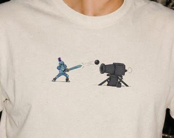 May include: A cream-colored short-sleeve t-shirt featuring a small graphic print on the chest. The illustration depicts a blue knight in armor holding a sword, facing a black cannon that is firing a round black cannonball toward the knight.
