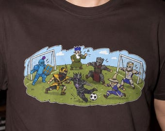 May include: A dark brown t-shirt featuring a graphic illustration of fantasy characters playing a soccer match on a green field. The scene includes a referee, two goal nets, and several armored figures in various poses, with one character kicking a soccer ball.
