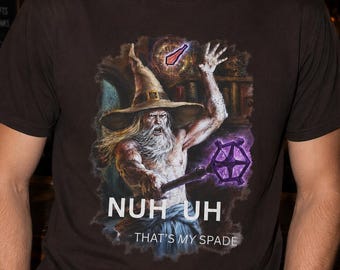 May include: A dark brown short-sleeve t-shirt featuring a graphic of a wizard with a long beard and hat, holding a purple glowing staff. Above the wizard is a small red spade icon. The text reads NUH UH THAT'S MY SPADE in white capital letters.