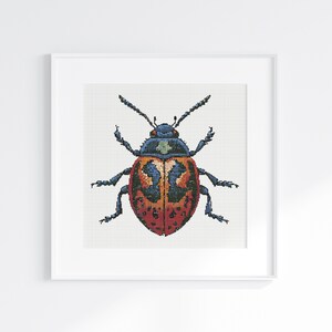 Op de afbeelding: Een ingelijste kruissteek van een kever. De kever heeft een rood lichaam met zwarte stippen, oranje en blauwe markeringen op de rug en blauwe poten en antennes. Het kunstwerk is in een witte lijst.
