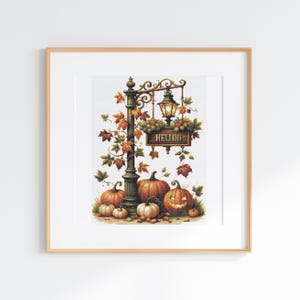 May include: A framed cross-stitch pattern featuring a dark green ornate lamppost adorned with autumn leaves and grapes. A wooden sign hanging from the post displays the text HELLOO. Several orange pumpkins sit at the base, including one jack-o'-lantern.