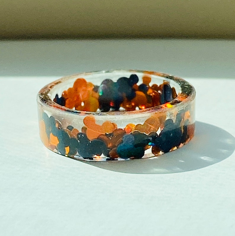 Puede incluir: Un anillo de resina transparente lleno de purpurina negra y naranja. La purpurina tiene forma de cabezas de Mickey Mouse.