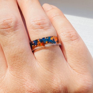 Puede incluir: Un anillo con una banda transparente y una capa de purpurina iridiscente en tonos azules, naranjas y dorados.