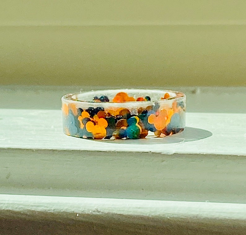 Puede incluir: Un anillo de resina transparente con un dise&ntilde;o colorido con cabezas de Mickey Mouse. El anillo est&aacute; lleno de cabezas de Mickey Mouse de color naranja, amarillo, azul, verde y negro.