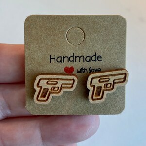 May include: A pair of wooden earrings shaped like handguns. The earrings are on a brown card with the text "Handmade with love".