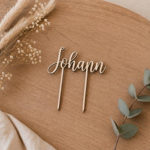 Op de afbeelding: Een taarttopper van lichtgekleurd hout met de naam Johann in een cursief lettertype. Het ontwerp bevat twee dunne, puntige stokjes die naar beneden uitsteken vanaf de letters, zodat het in een taart of andere ondergrond gestoken kan worden.