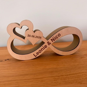 May include: A light-colored wooden infinity symbol ornament featuring two integrated heart shapes. The surface is engraved with the text 'Leonie & Nico' and the date '20.06.2026' in a dark brown font, creating a decorative piece with smooth edges.