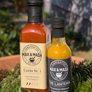 Könnte beinhalten: Zwei Glasflaschen handgemachter Chilisauce, "Cuvée Nr. 1" und "The Lantern", mit schwarzen Verschlüssen und Etiketten. Die Etiketten tragen den Markennamen "MAX & MASH" und den Text "EST. 2026". Die Flaschen stehen vor einem Hintergrund aus grünen Pflanzen.