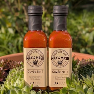 Könnte beinhalten: Zwei kleine Glasflaschen mit roter Chilisauce und schwarzen Verschlüssen. Jede Flasche trägt ein Etikett mit der Aufschrift "MAX & MASH" und "Cuvée Nr. 1". Die Flaschen sind von grünen und roten Sukkulenten umgeben.