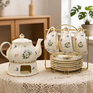 May include: A white ceramic tea set featuring a teapot on a warmer base and three teacups hanging on a gold metal rack above a stack of matching saucers. Each piece is decorated with a delicate floral pattern of blue, pink, and green flowers.