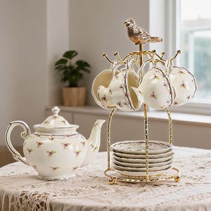 Peut inclure: Une théière en porcelaine crème et un ensemble assorti de quatre tasses et soucoupes. Les pièces présentent un délicat motif floral rose et une bordure dorée. Les tasses sont suspendues à un support en métal doré surmonté d'un oiseau.