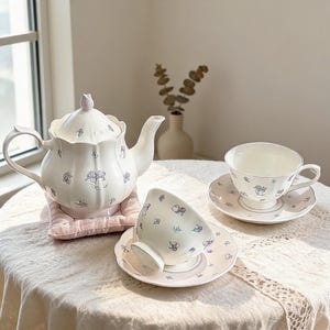 Puede incluir: Una tetera de cerámica blanca con tapa festoneada y tazas a juego sobre platillos, decoradas con pequeños motivos florales morados. La tetera descansa sobre un posavasos acolchado de color rosa claro, con un diseño delicado y redondeado.