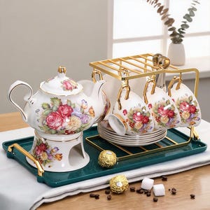 Pode incluir: Um conjunto de chá em porcelana branca com um padrão floral de rosas vermelhas, amarelas e cor-de-rosa. O conjunto inclui um bule sobre uma base de aquecimento, chávenas e pires, dispostos num tabuleiro retangular verde-escuro com suporte.