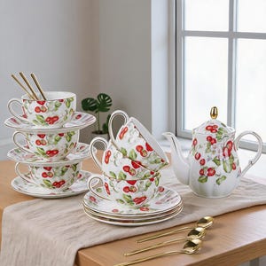 Puede incluir: Un juego de té de porcelana blanca con un patrón de cerezas rojas y hojas verdes con bordes dorados. El conjunto incluye una tetera con remate dorado, varias tazas con sus platos a juego y cucharas delgadas de tono dorado.