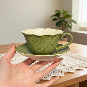 May include: A ceramic teacup and saucer set featuring a textured, mottled green exterior that mimics the appearance of cabbage leaves. The cup has a curved handle and a scalloped rim, while the saucer has a matching leaf-shaped design.