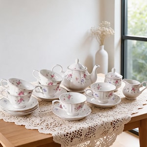 Puede incluir: Un juego de té de porcelana blanca con un delicado patrón de rosas rosas y flores azules con bordes dorados. El conjunto incluye una tetera con tapa, un azucarero con tapa, una jarra de leche y cuatro tazas con sus platos a juego.