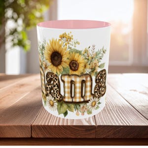 Könnte beinhalten: Weißer Keramikbecher mit rosa Innenraum, mit dem Wort "Mama" in einer Mischung aus Leopardenmuster und gelbem Vichy-Karo. Der Becher ist mit Sonnenblumen, Gänseblümchen und anderen floralen Elementen verziert.