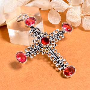 May include: A silver-colored cross pendant with five oval, red gemstones. The cross has an ornate, filigree design. The pendant is displayed on a light orange surface with white flowers in the background. The cross is a religious symbol.