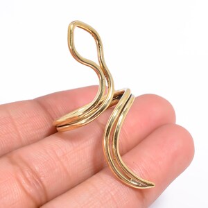 May include: A gold-toned metal ring featuring a double-wire design that wraps around the finger. The ring has an open, elongated shape with two distinct curved sections that extend vertically, creating a continuous, fluid, and polished metallic look.