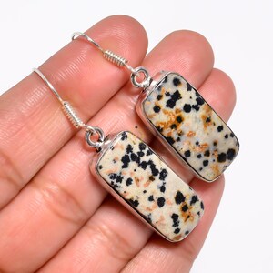 May include: A pair of rectangular earrings with a speckled, cream-colored stone and black and brown spots. The stone is set in a silver-colored metal frame and hangs from a silver-colored hook.