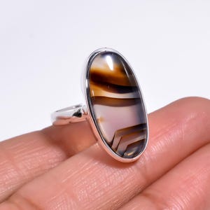 May include: A silver ring featuring a large, oval-shaped cabochon gemstone with horizontal bands of translucent white, light brown, and dark brown colors. The stone is set in a smooth, polished silver bezel with a simple, slender silver band.