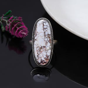 Può includere: Anello ovale in metallo argentato con una pietra bianca venata di marrone. La pietra è incorniciata da un bordo a corda intrecciata. Il design dell'anello è semplice ma elegante, adatto all'uso quotidiano o alle occasioni speciali.
