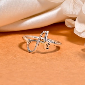 May include: A silver-colored ring featuring the letter "A" in a cursive font. The ring is set against a warm, orange-toned background, with a soft, white fabric in the background. The ring is a piece of jewelry.