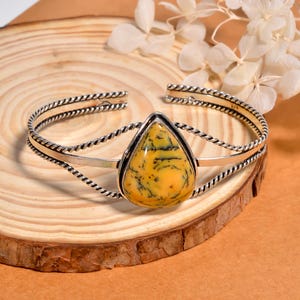 Könnte beinhalten: Silbernes Armreif-Armband mit einem tropfenförmigen, gelb-schwarzen Stein. Das Armband hat ein gedrehtes Seildesign an den Kanten und ein poliertes Silberband. Der Stein ist in einer silbernen Lünette gefasst.
