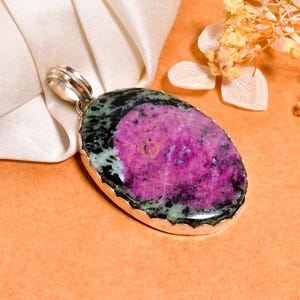 Peut inclure: Un pendentif ovale composé d'une pierre précieuse de rubis zoïsite polie, présentant un centre magenta vif entouré d'inclusions minérales vert foncé et noires, serti dans une monture en métal argenté festonnée avec une bélière circulaire.