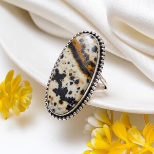 May include: An oval-shaped ring featuring a polished dalmatian jasper stone with black and tan spots set in a sterling silver bezel. The metal setting is decorated with a beaded texture along the outer edge, creating a detailed frame for the stone.