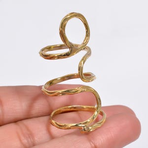 May include: A gold-toned metal finger splint ring featuring a continuous, spiraling design with an organic, textured surface. The structure consists of three interconnected circular loops that wrap around the finger, providing a layered appearance.