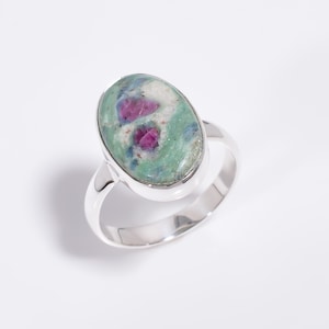 May include: A silver ring featuring an oval-shaped cabochon stone. The stone displays a marbled pattern with prominent inclusions of vibrant magenta-pink, interspersed with shades of light green, white, and subtle dark blue mineral veins.