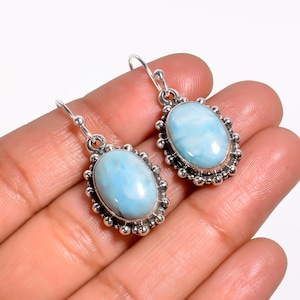 May include: A pair of silver drop earrings featuring oval, light blue gemstones. The gemstones are set within a decorative, beaded silver frame. The earrings are displayed against a neutral background.