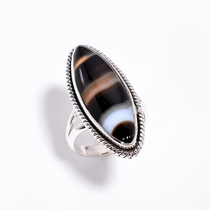 May include: A silver ring featuring a large, elongated marquise-shaped stone with horizontal bands of black, white, and translucent brown. The stone is set in a polished silver bezel surrounded by a decorative twisted rope-style metal border.