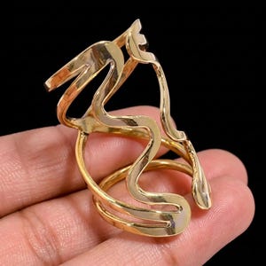 May include: A gold-toned metal ring featuring an open, multi-layered design with fluid, wavy lines that wrap around the finger. The structure consists of several interconnected, polished bands that create a continuous, abstract, and elongated silhouette.